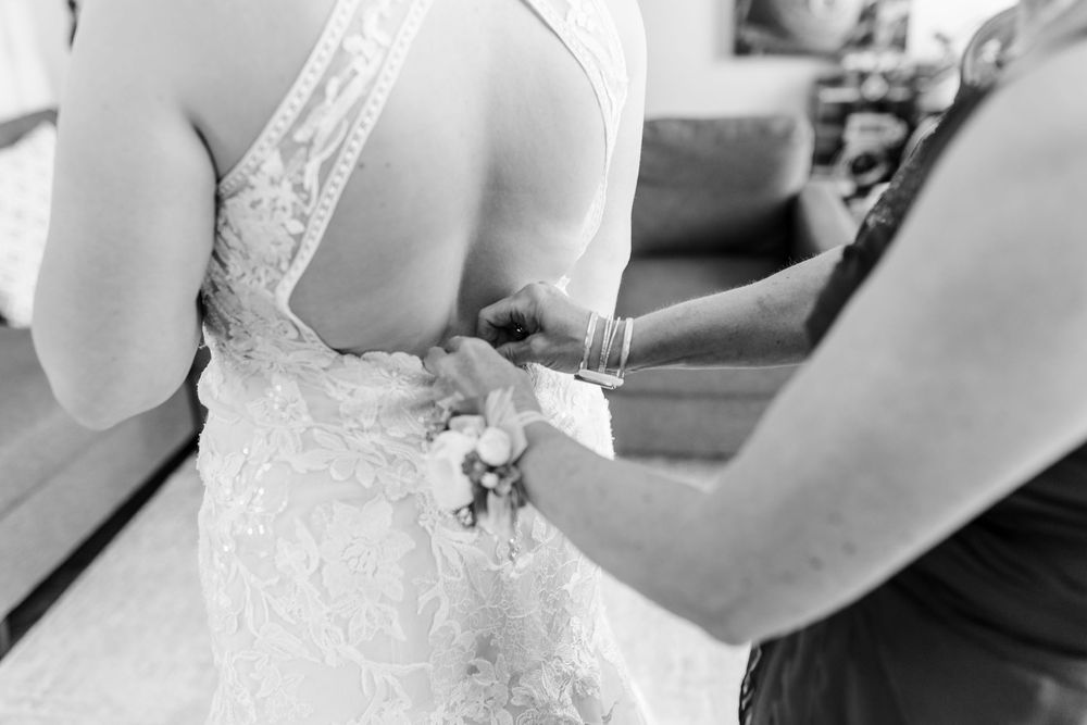 Black and white image of a bride in a lace dress being helped with final wedding preparations.
