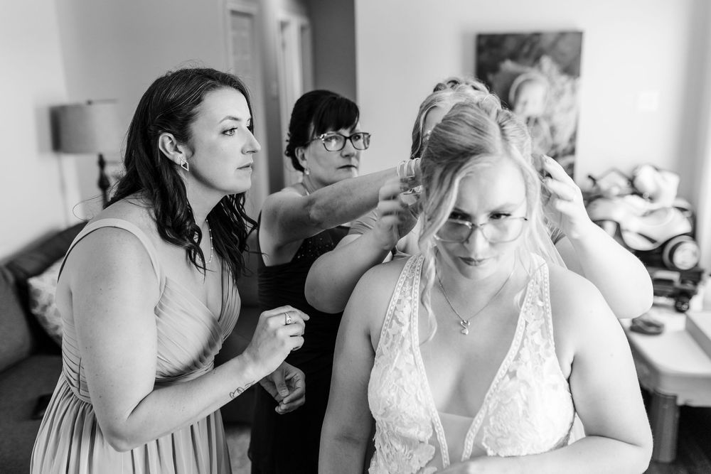 Black and white photo showing wedding preparations with bridesmaids helping style a bride's hair in a getting ready room.