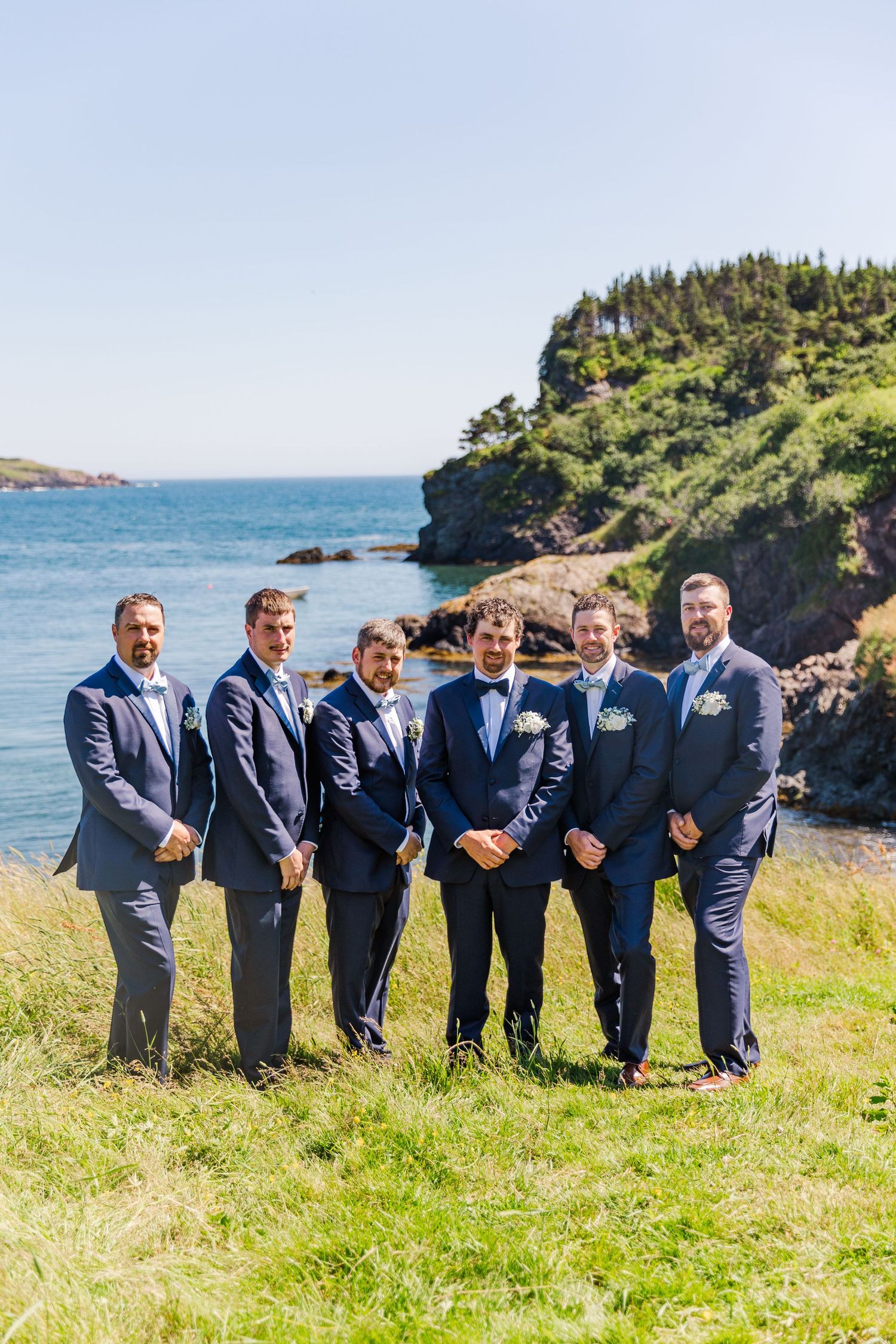 A wedding party in navy suits stands on a grassy cliff overlooking the blue ocean on a sunny day.