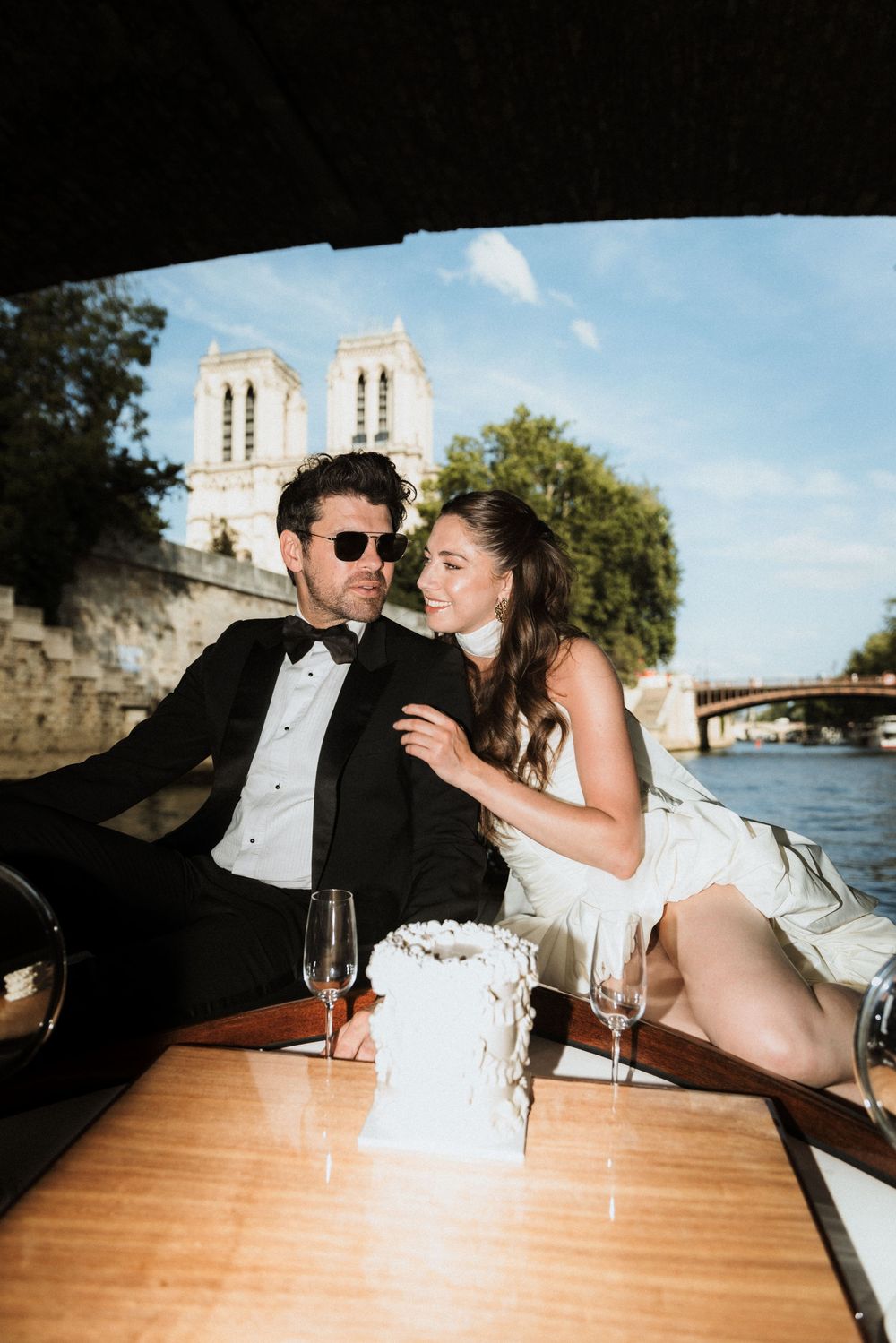 A couple in formal attire share a romantic moment at an outdoor venue with wine glasses on a wooden table.
