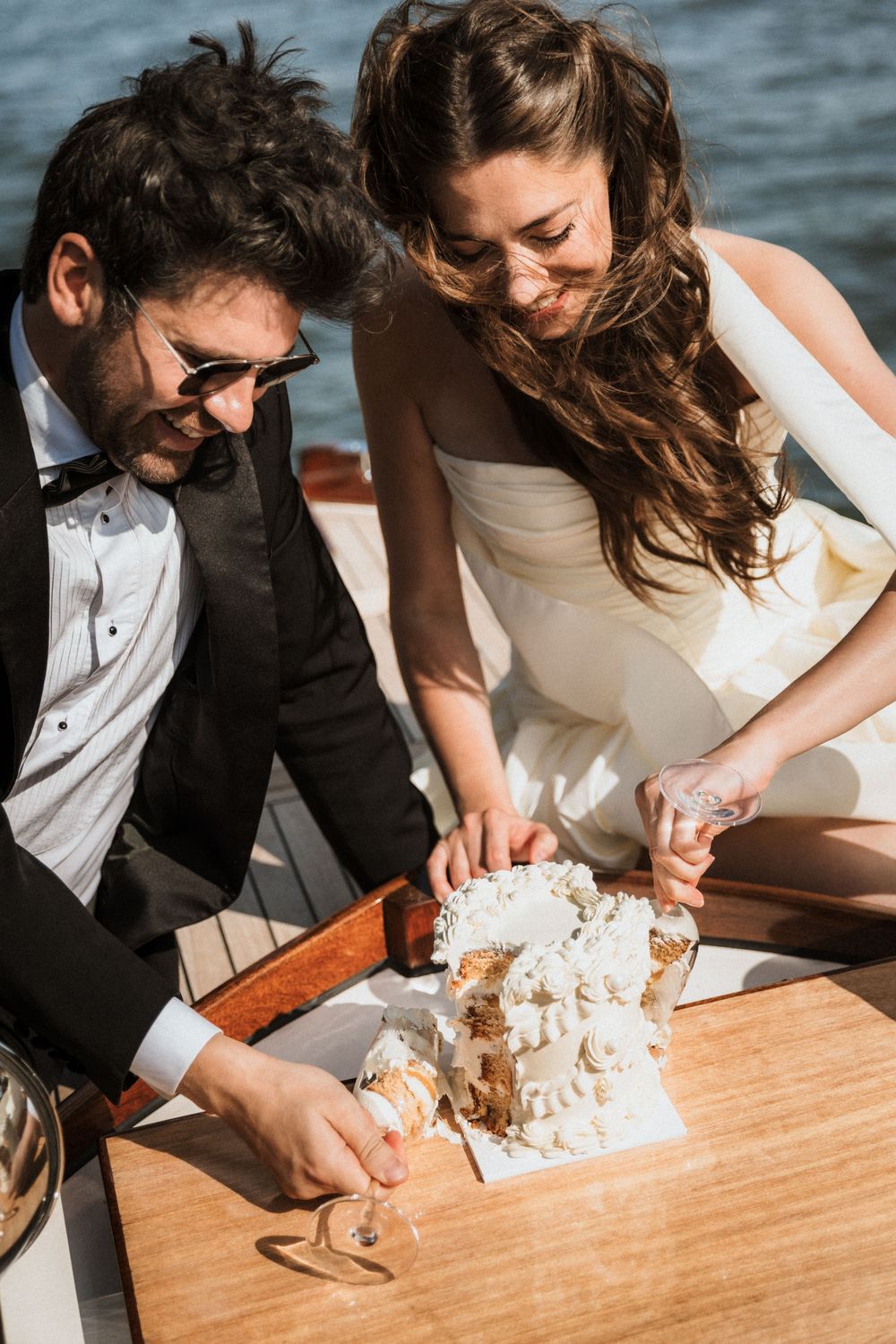 A couple in wedding attire cuts into a white cake together at a wooden table.