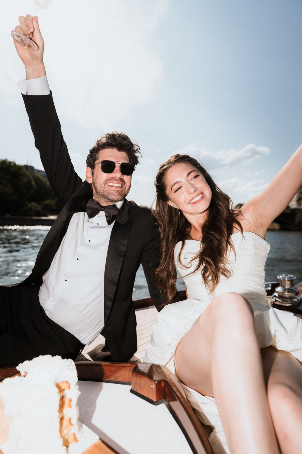 A newlywed couple celebrates joyfully on a boat during their wedding day, with sunglasses and formal attire.