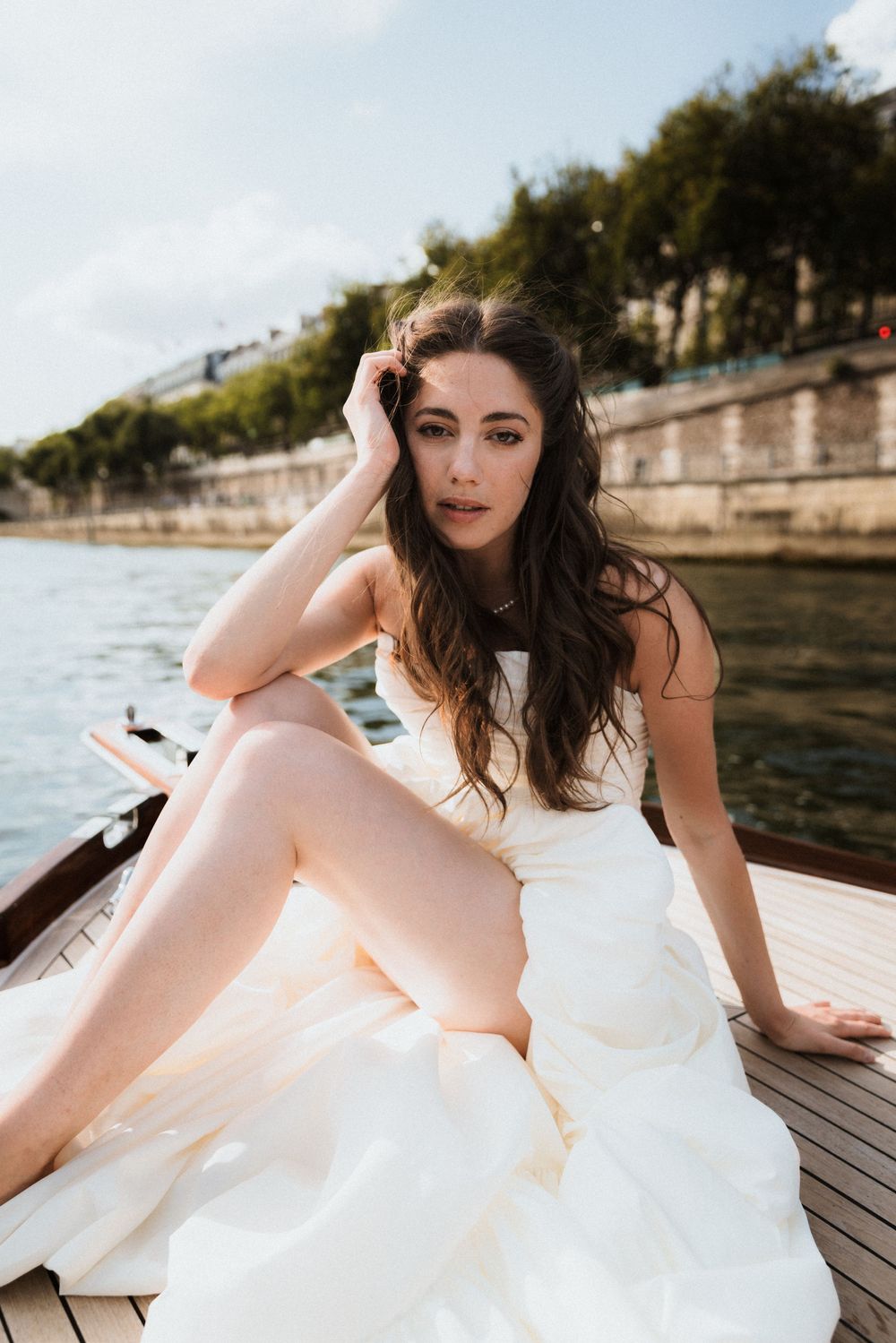 A person in a flowing white dress poses on a wooden deck by a waterfront on a sunny day.
