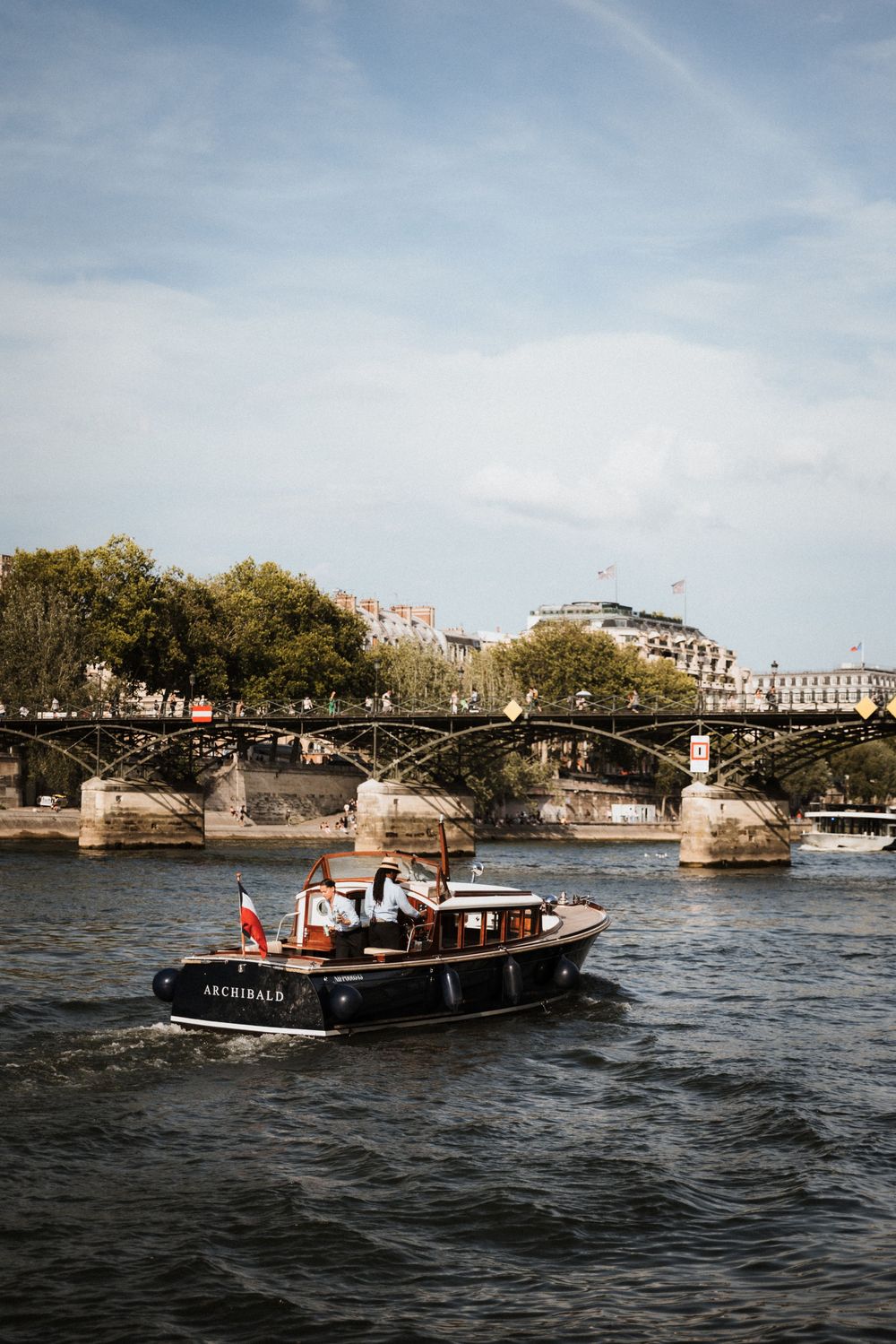 A small boat cruises on the Seine River in Paris with the historic Pont des Arts bridge in the background.
