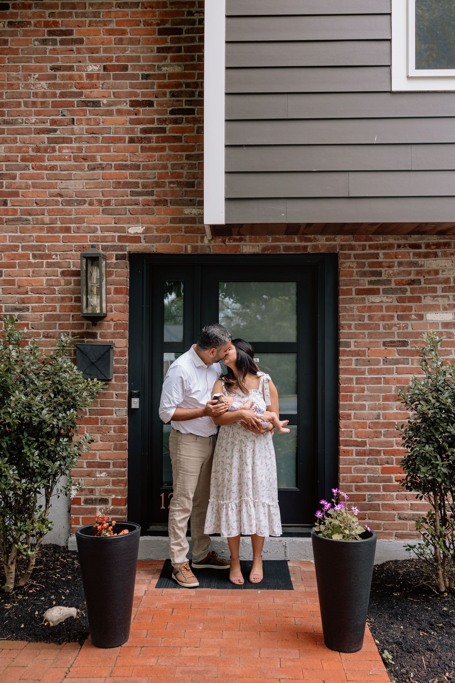 A couple stands together on a brick porch with black planters and a glass door entrance of their home.