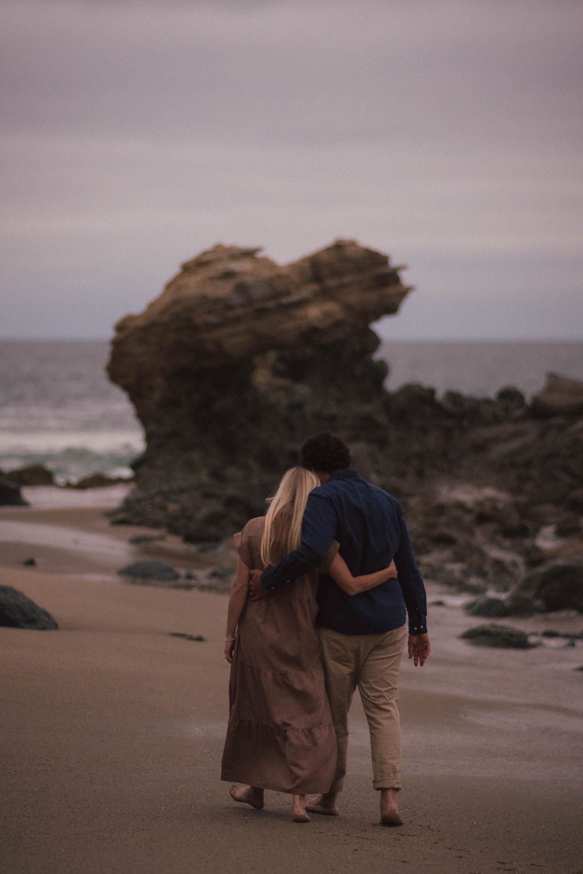 A couple walks intimately along a moody beach with dramatic rock formations at sunset.