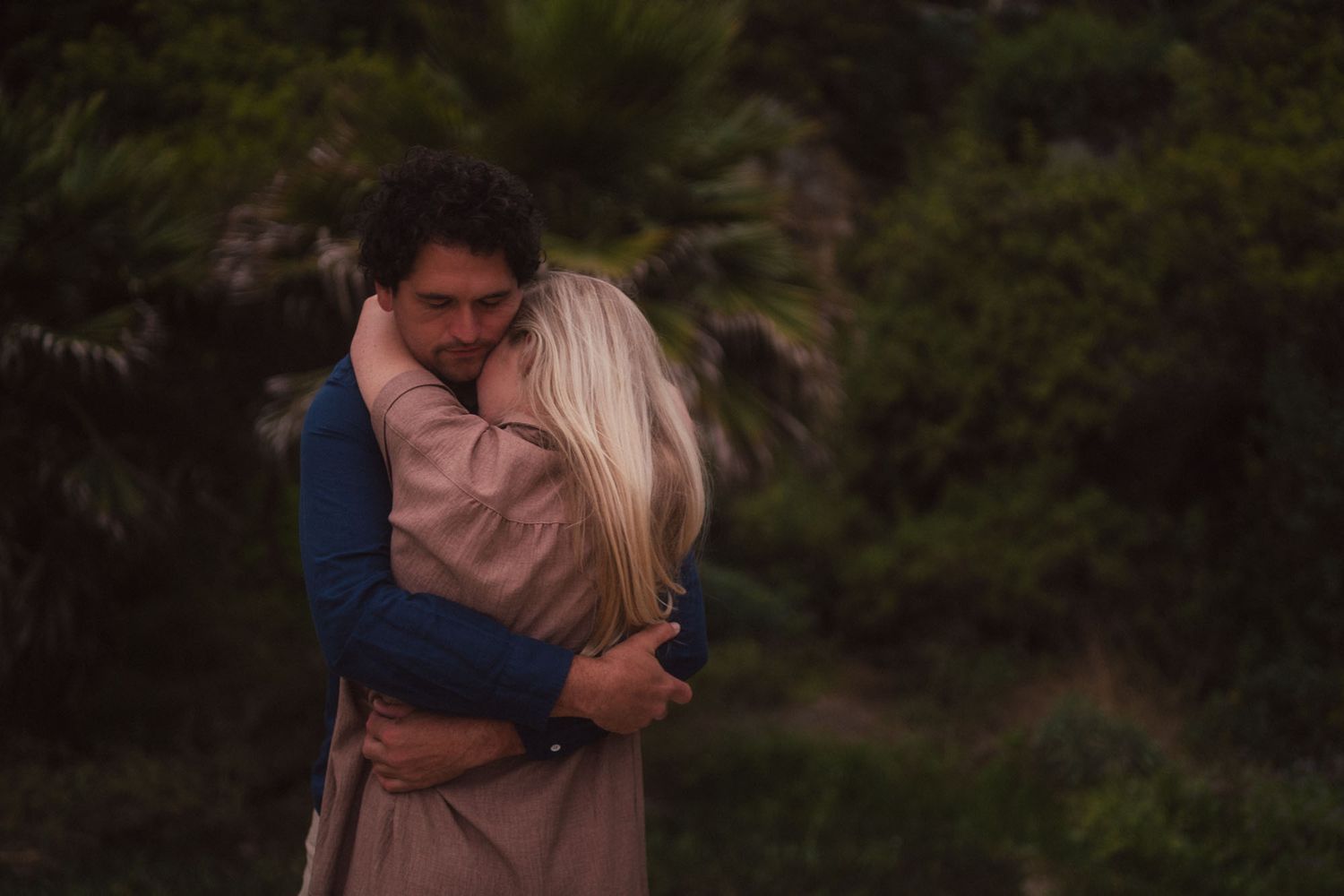 A couple embraces tenderly in a romantic outdoor setting surrounded by lush greenery and natural light.