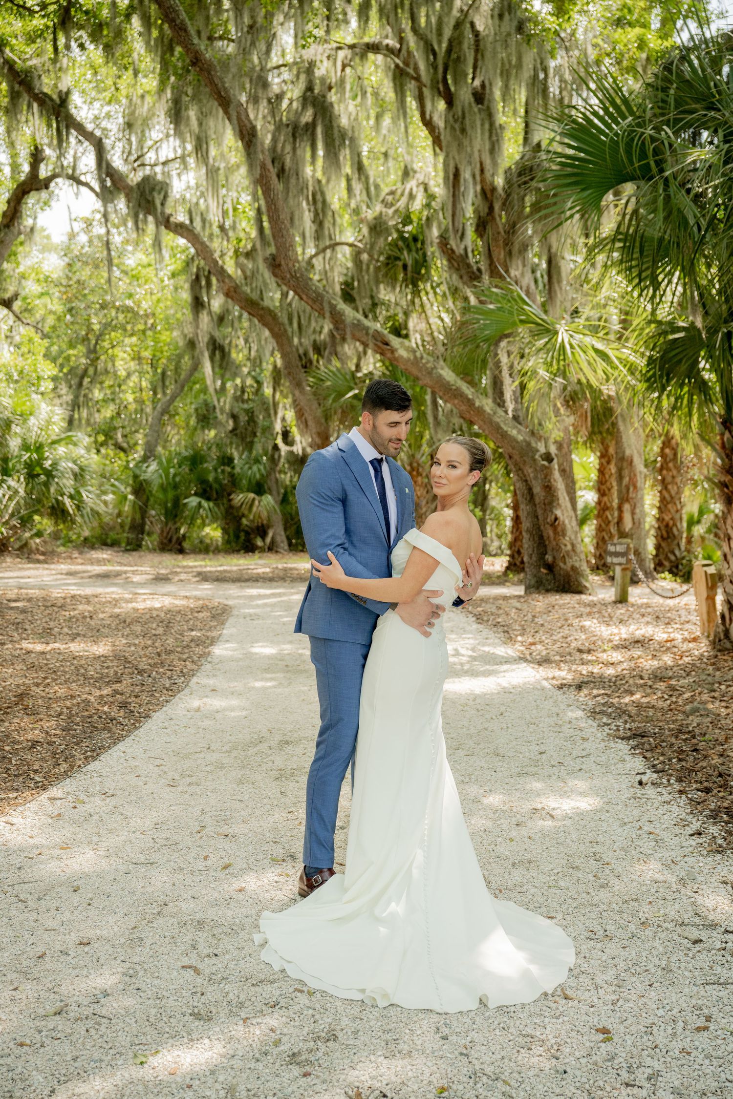 A couple shares a romantic moment on a path lined with Spanish moss-draped oak trees in a dreamy outdoor setting.