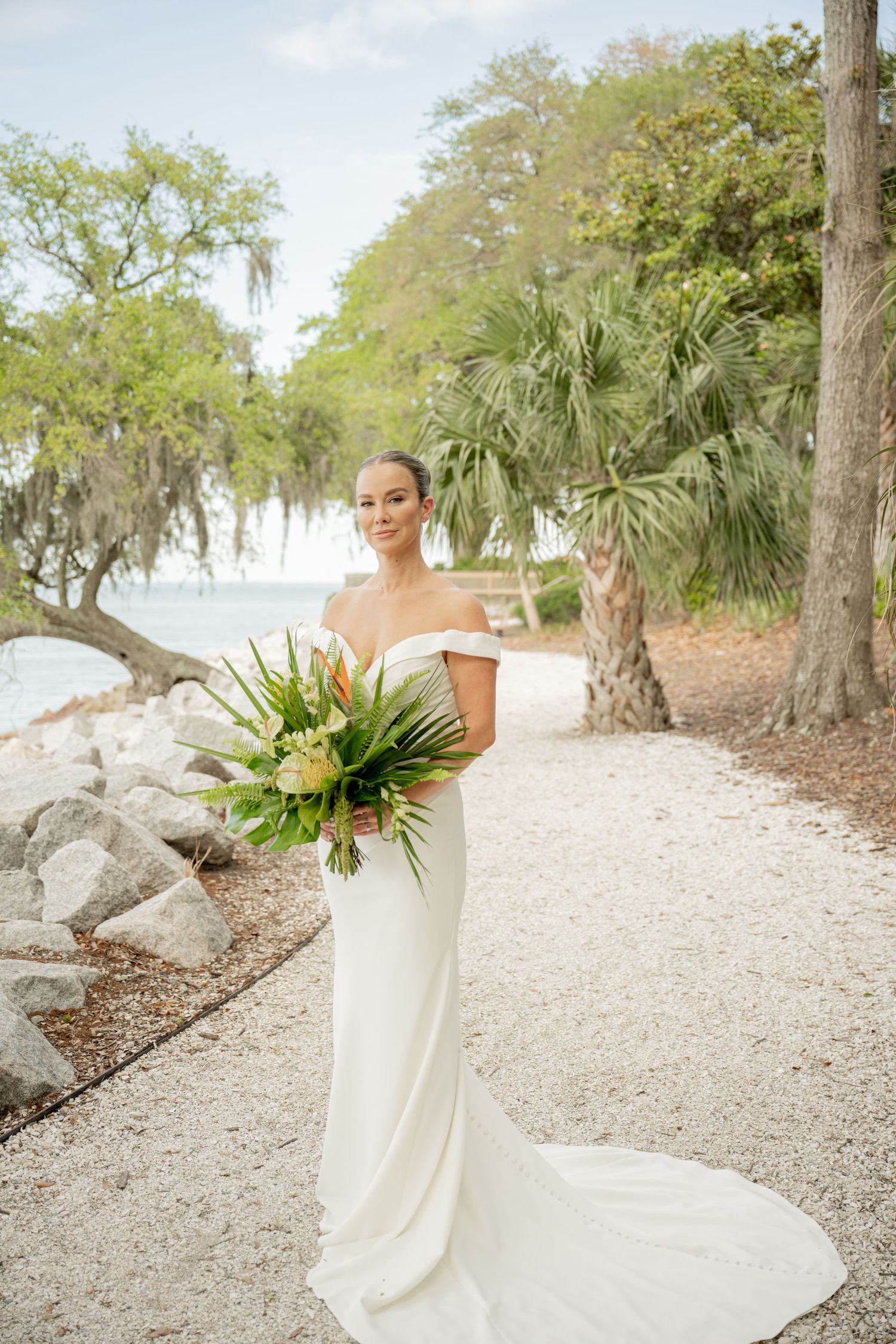 Elegant off-shoulder wedding dress with tropical bouquet on scenic coastal path lined with palm trees and Spanish moss.