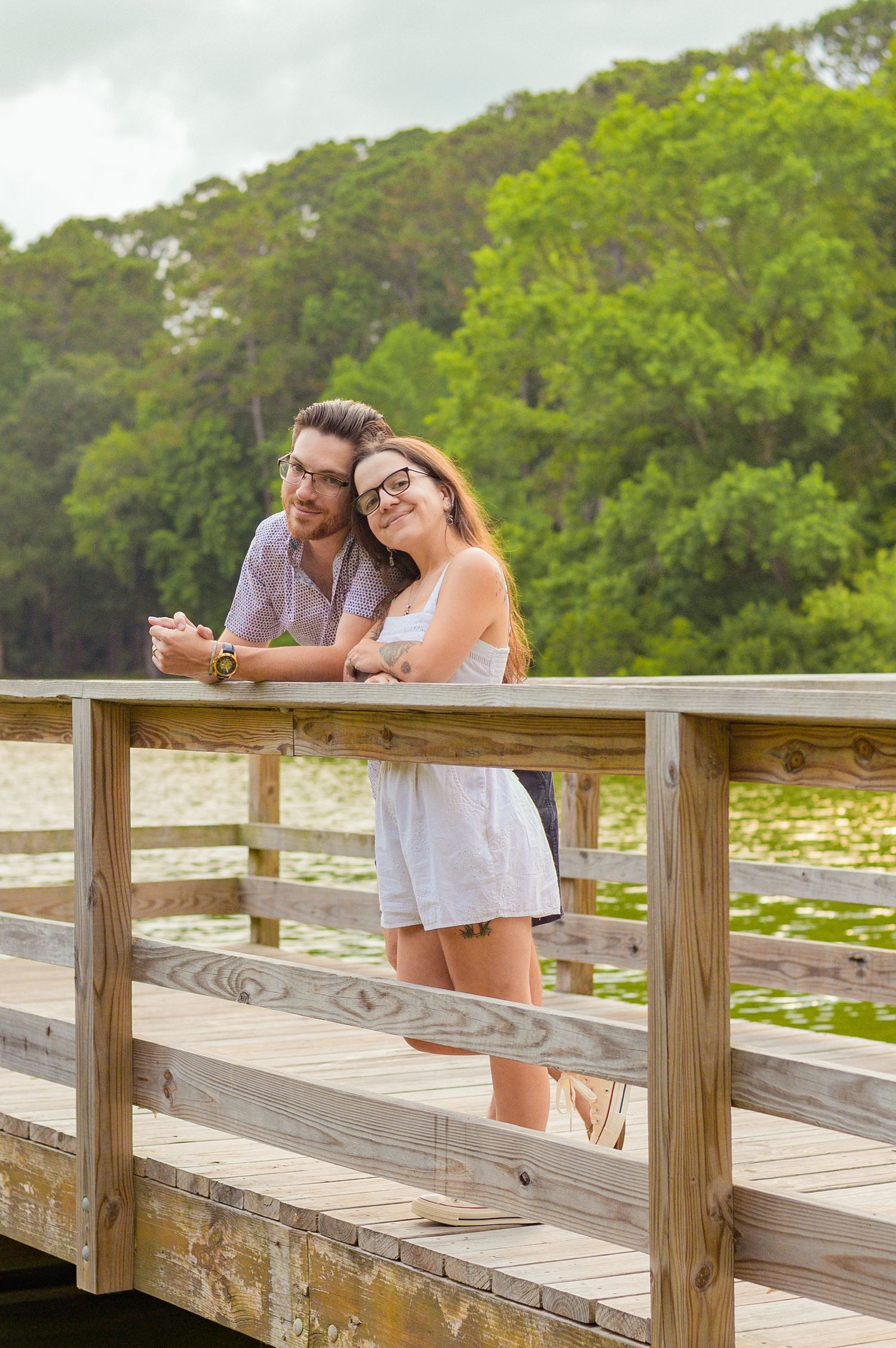 A couple sharing a happy moment while leaning on a wooden dock railing surrounded by trees.