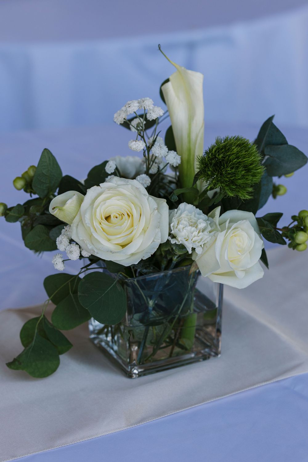 Elegant white roses and calla lily arranged in a square glass vase with green accents against a soft blue background.