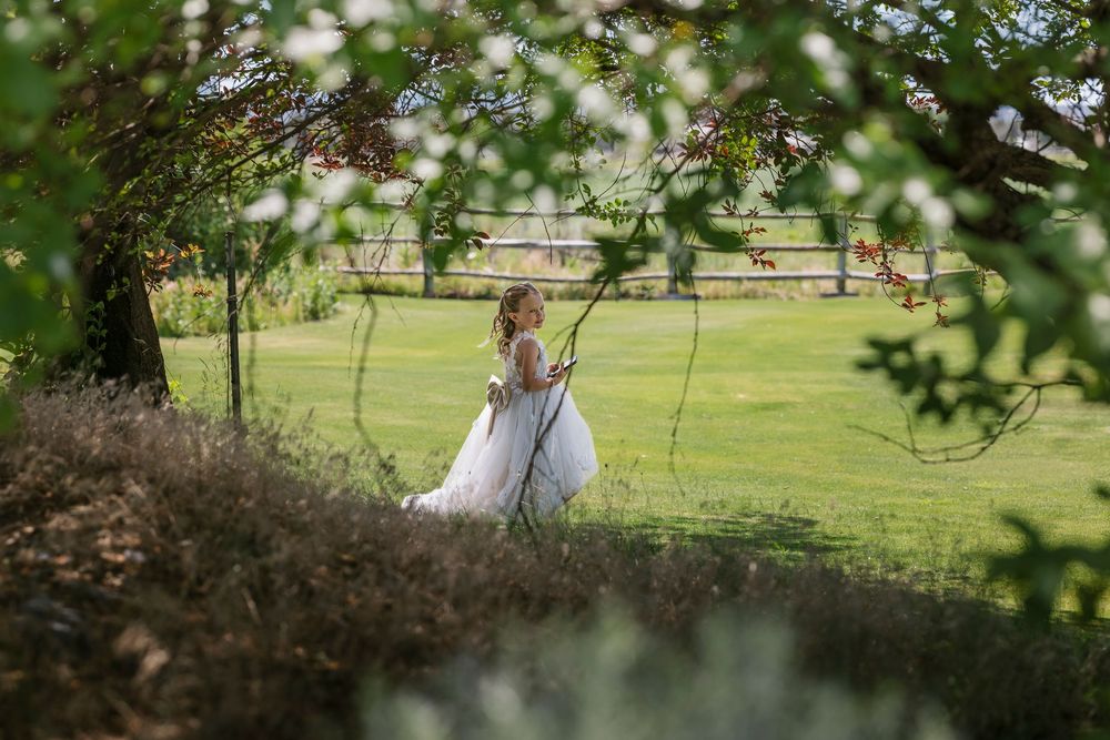 A bride in a white wedding gown sits on a rustic wooden swing in a sunlit garden setting.