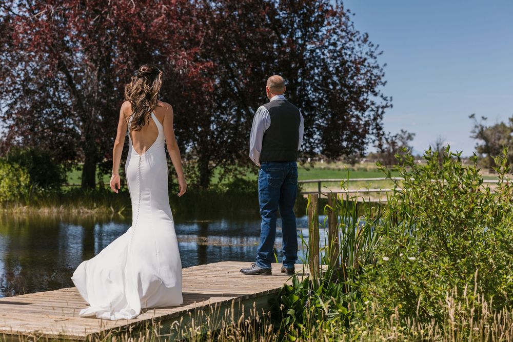 A couple stands on a wooden dock overlooking a tranquil pond surrounded by green foliage on a sunny summer day.