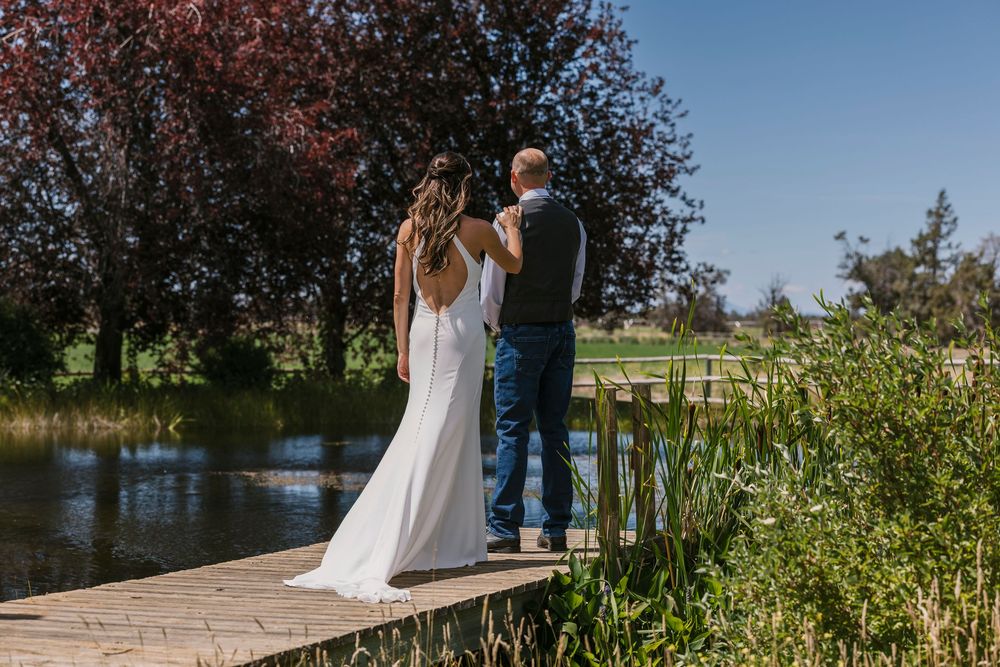 A couple stands together during an outdoor photo session near a scenic pond surrounded by lush greenery and pink florals.
