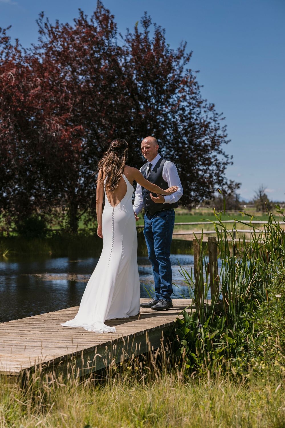 A couple shares a tender moment on a wooden dock surrounded by tall grass and trees near a lake on their wedding day.