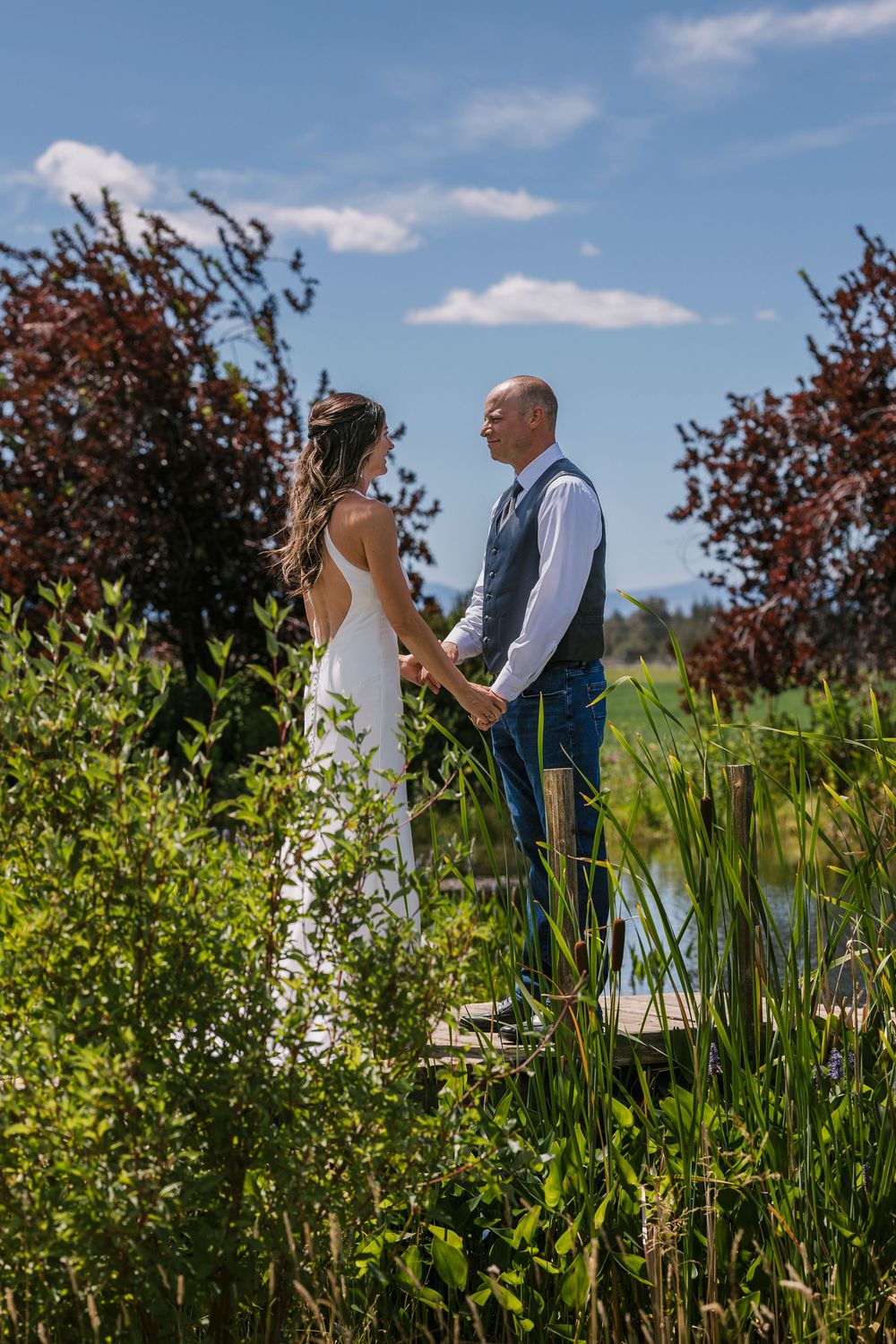 A wedding couple stands together among vibrant green grasses and wildflowers on a sunny summer day.