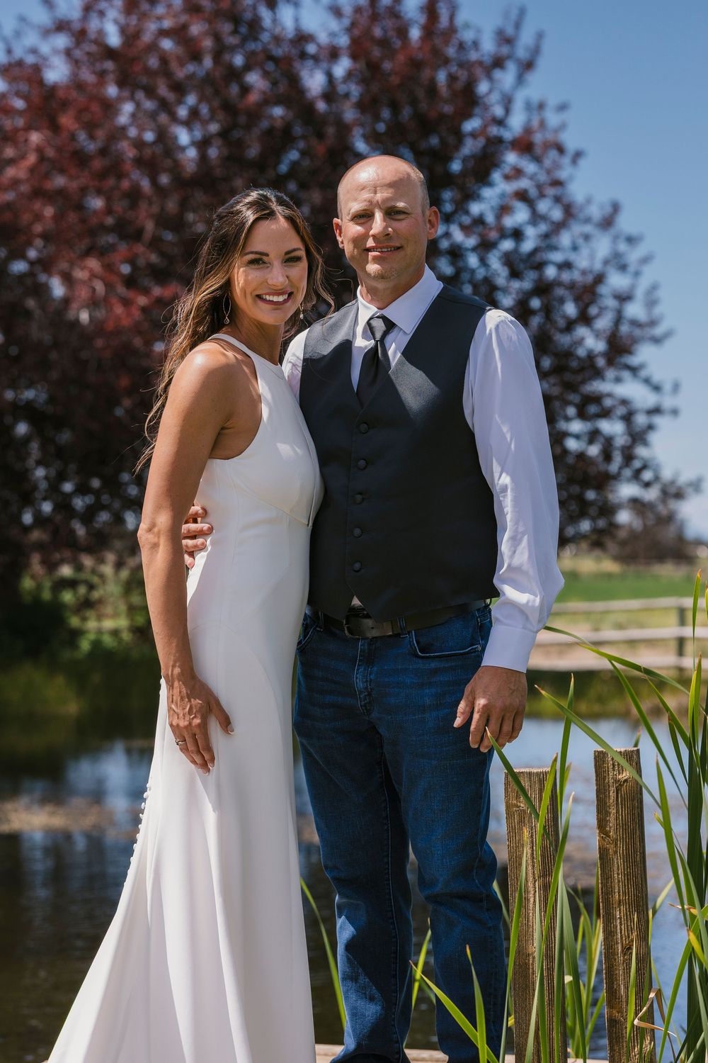 A couple stands together by a pond on their wedding day against flowering trees in the background.
