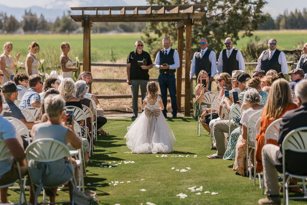 Outdoor wedding ceremony takes place under wooden pergola on green lawn with guests seated in white chairs on sunny day.