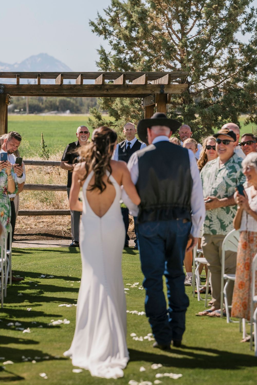 Wedding ceremony guests gather outdoors under blue skies with mountains visible in the background.