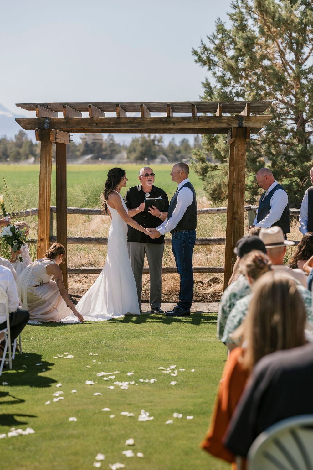 Outdoor wedding ceremony under a wooden pergola on a grassy lawn with guests watching on a sunny day.