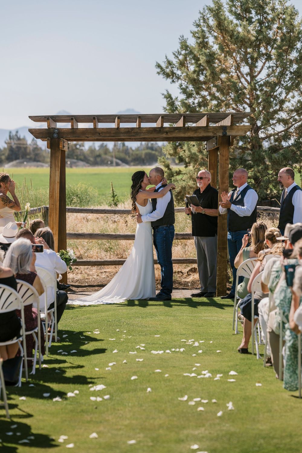 A beautiful outdoor wedding ceremony under a wooden pergola with guests seated on white chairs on a grassy lawn.