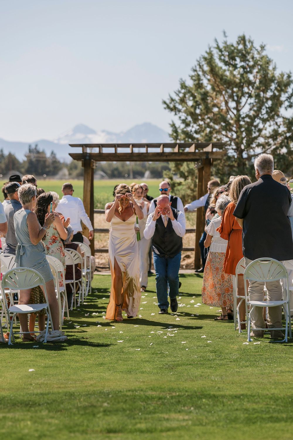 Wedding guests gather on a manicured lawn with mountains visible in the background during an outdoor ceremony.