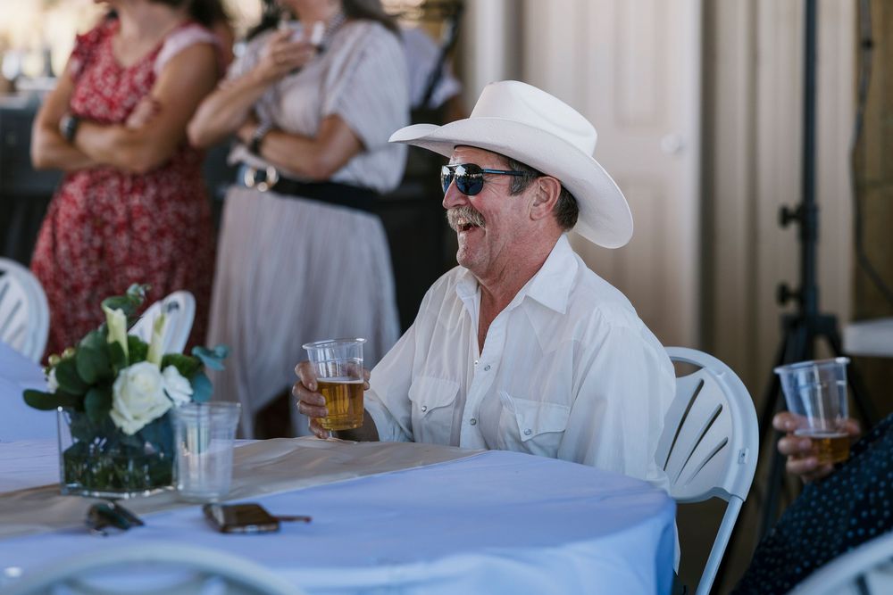 Guest in a white cowboy hat and sunglasses enjoys a drink at an outdoor event with table settings.