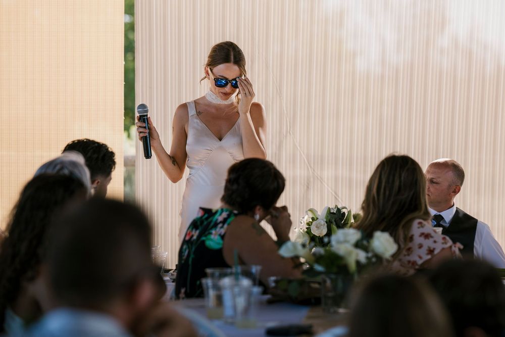 Wedding guests at a long table celebrate during an outdoor reception with floral decorations and champagne glasses.