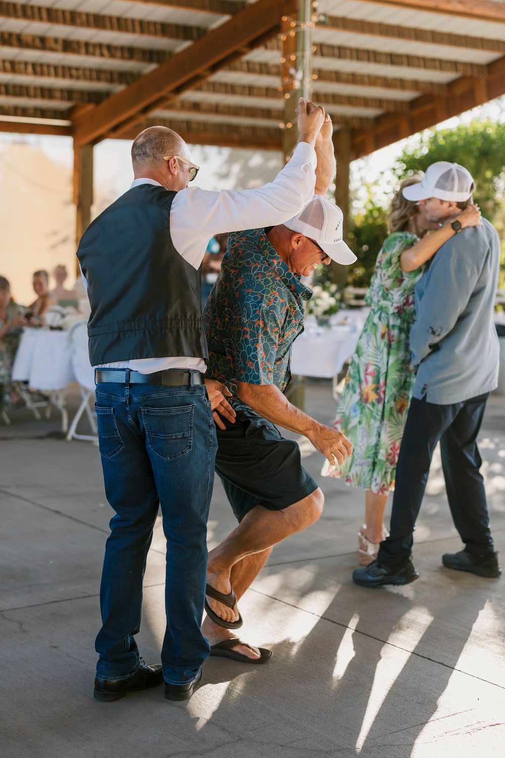 Wedding guests dance together under a covered patio, casting long shadows in the afternoon light.