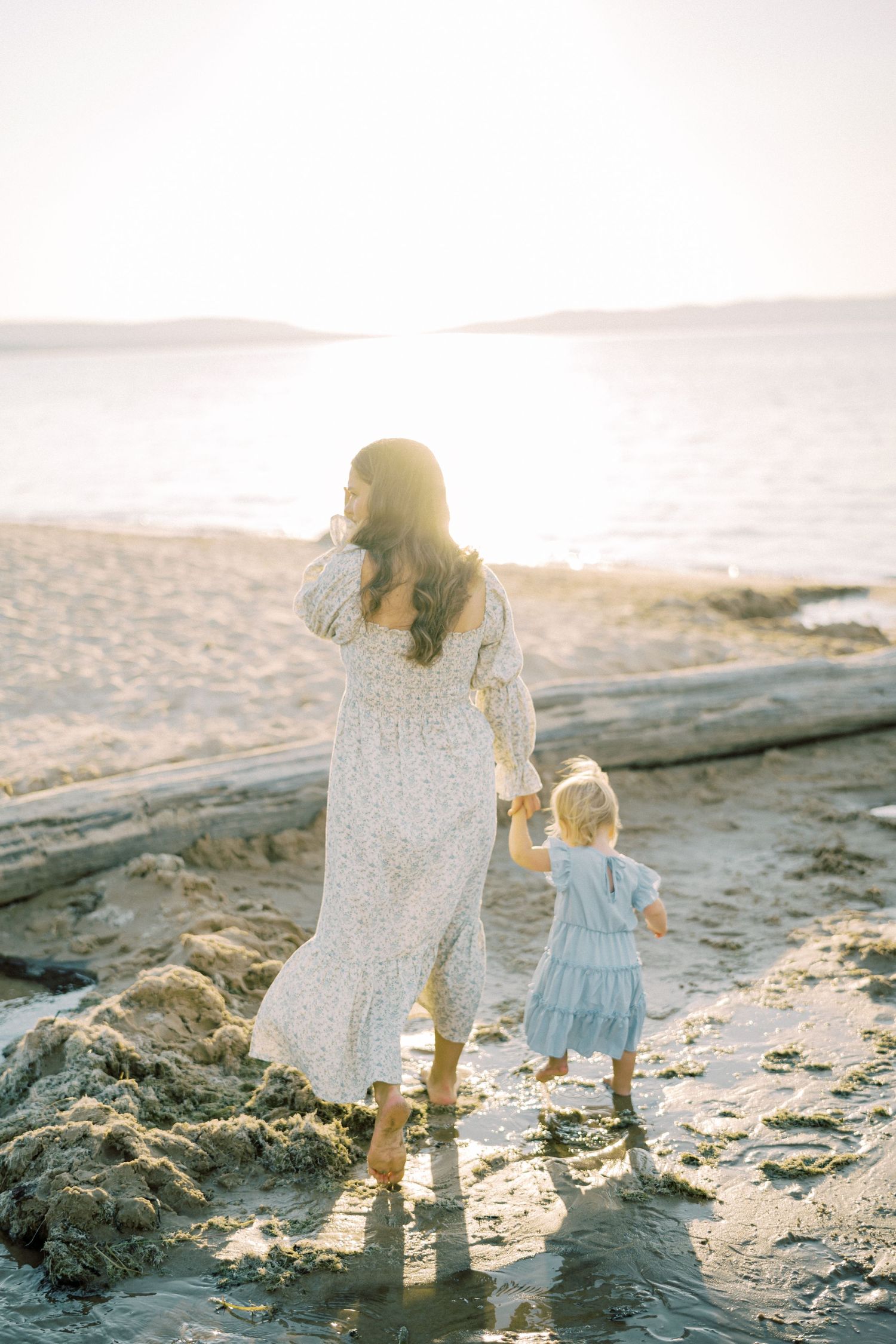 Mother and child walking along beach shore at sunset in flowing dresses.