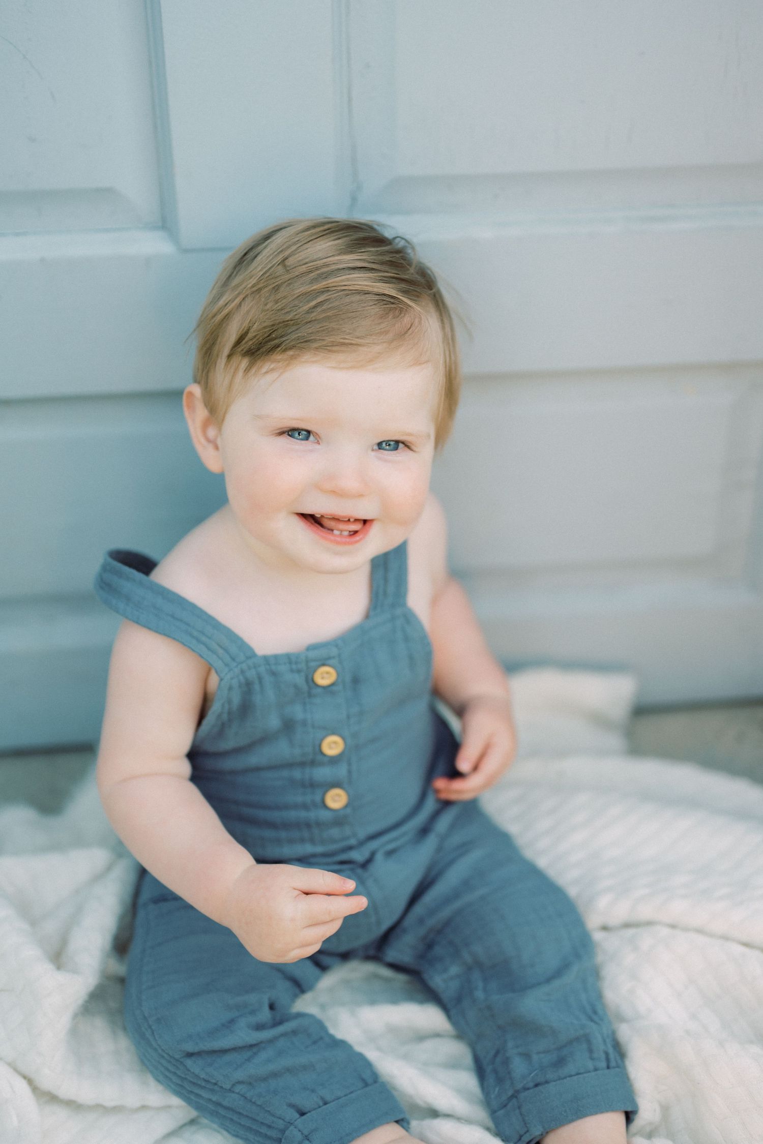 Adorable toddler wearing blue denim overalls smiles while sitting on a white blanket against a blue wall.
