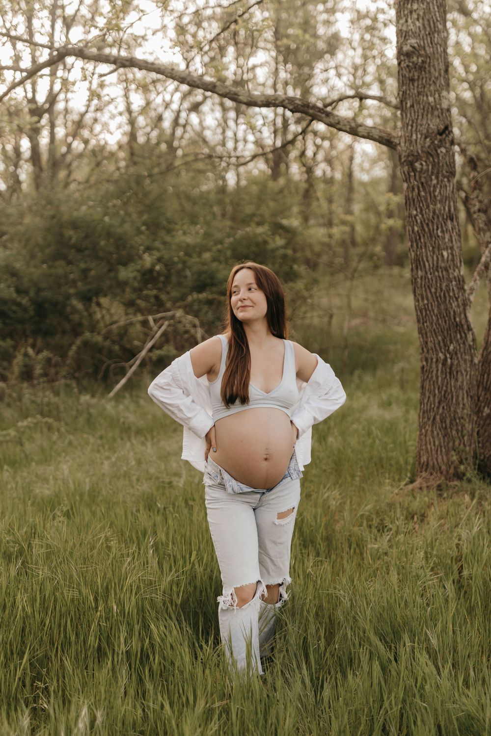 A solo maternity portrait in a white outfit standing among tall grass and trees.