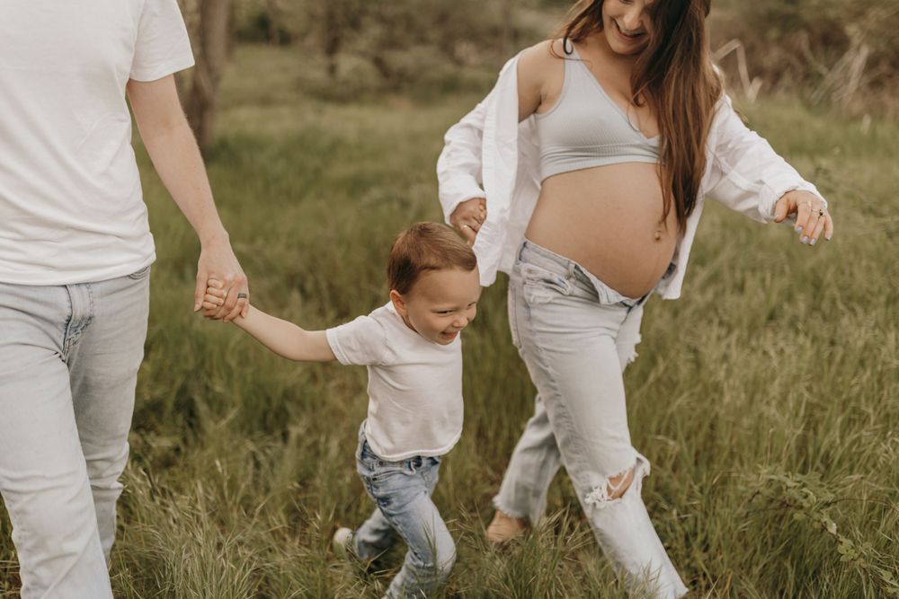 Family walks together in a grassy field with a pregnant woman and toddler holding hands.
