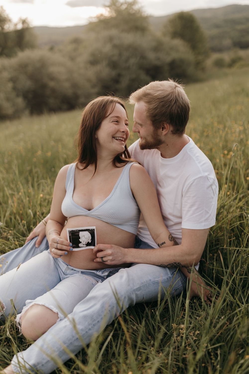 A couple shares a happy moment while holding an ultrasound photo in a grassy field.