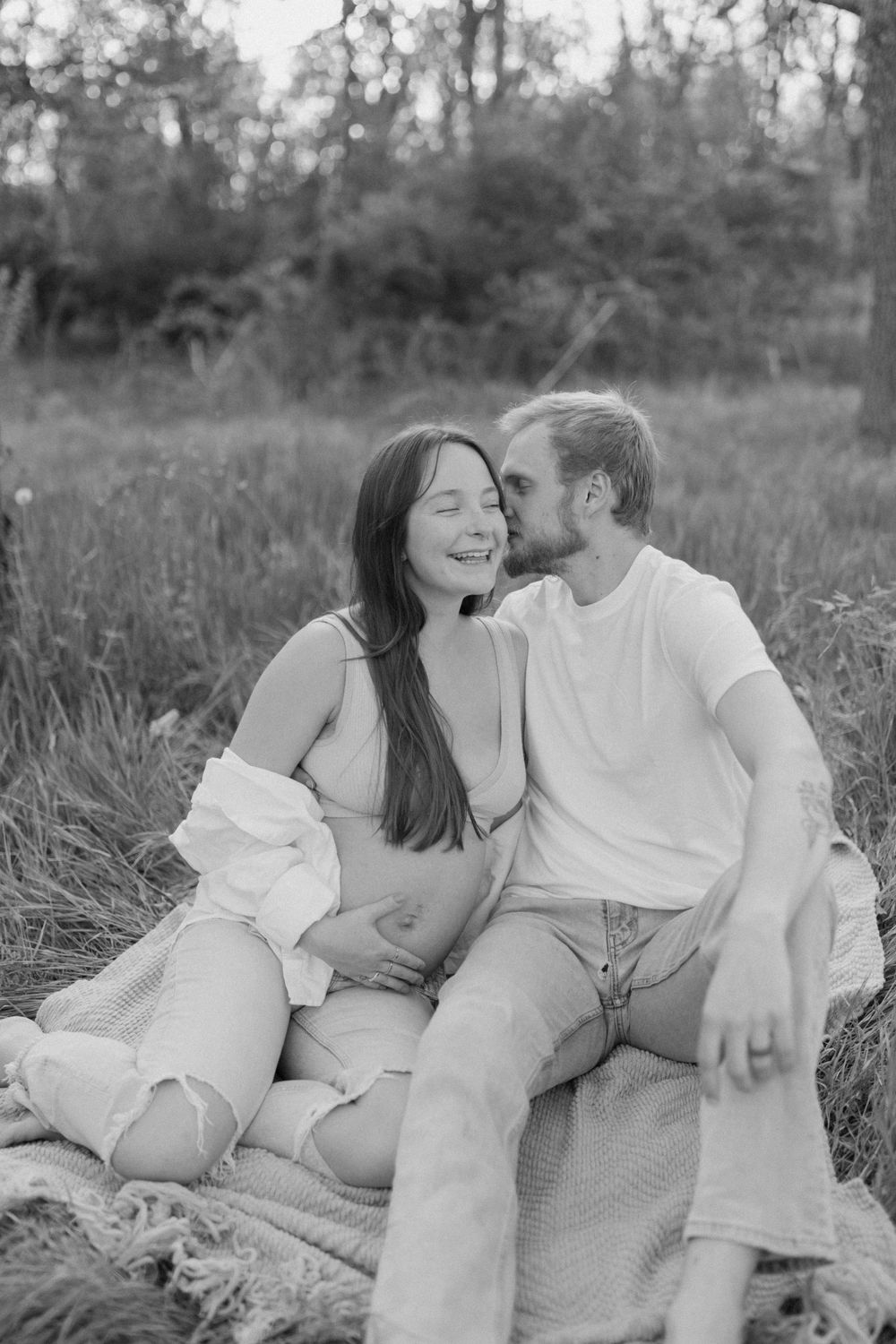 A black and white outdoor maternity photo of a couple sharing a joyful moment on a picnic blanket.