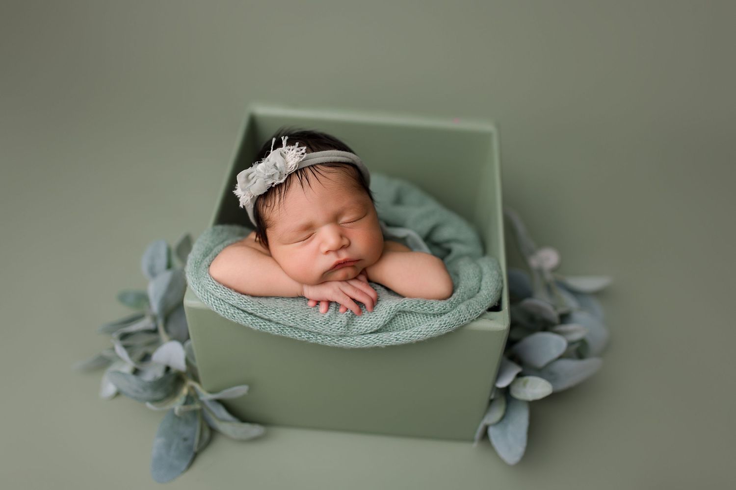A sleeping newborn wearing a white headband rests peacefully in a sage green prop bowl with soft fabric decorations.
