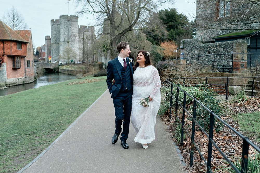 A couple in wedding attire walk alongside ancient stone walls and castle ruins on a winter day in England.