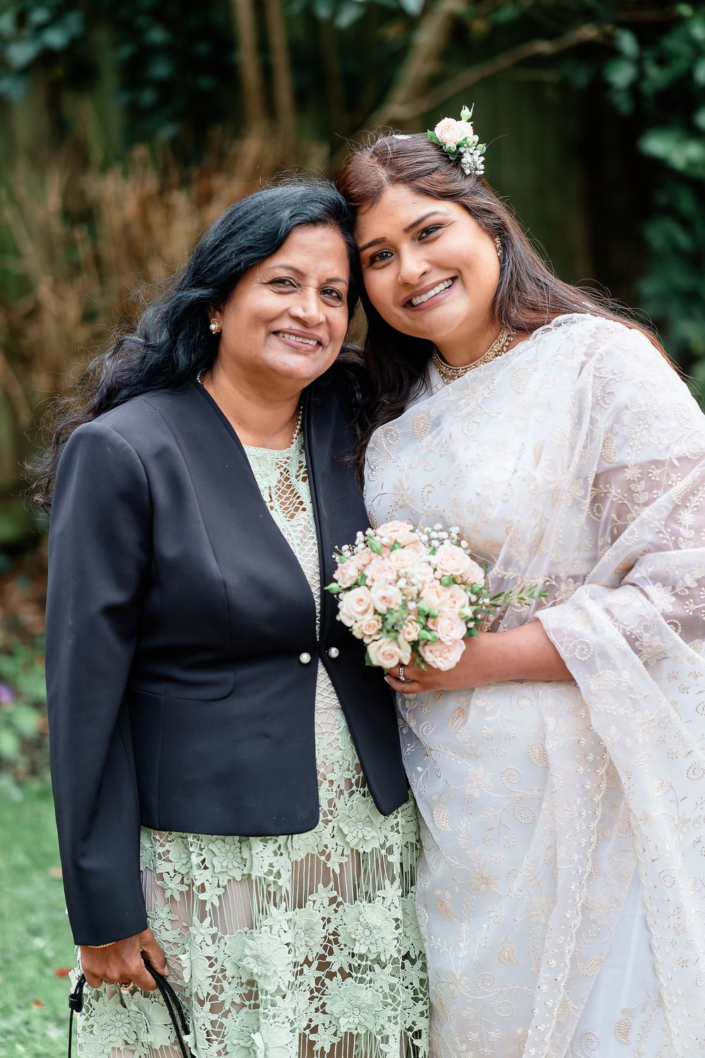 Two people wearing formal attire outdoors sharing a moment with a bridal bouquet and pearl white outfits against greenery.