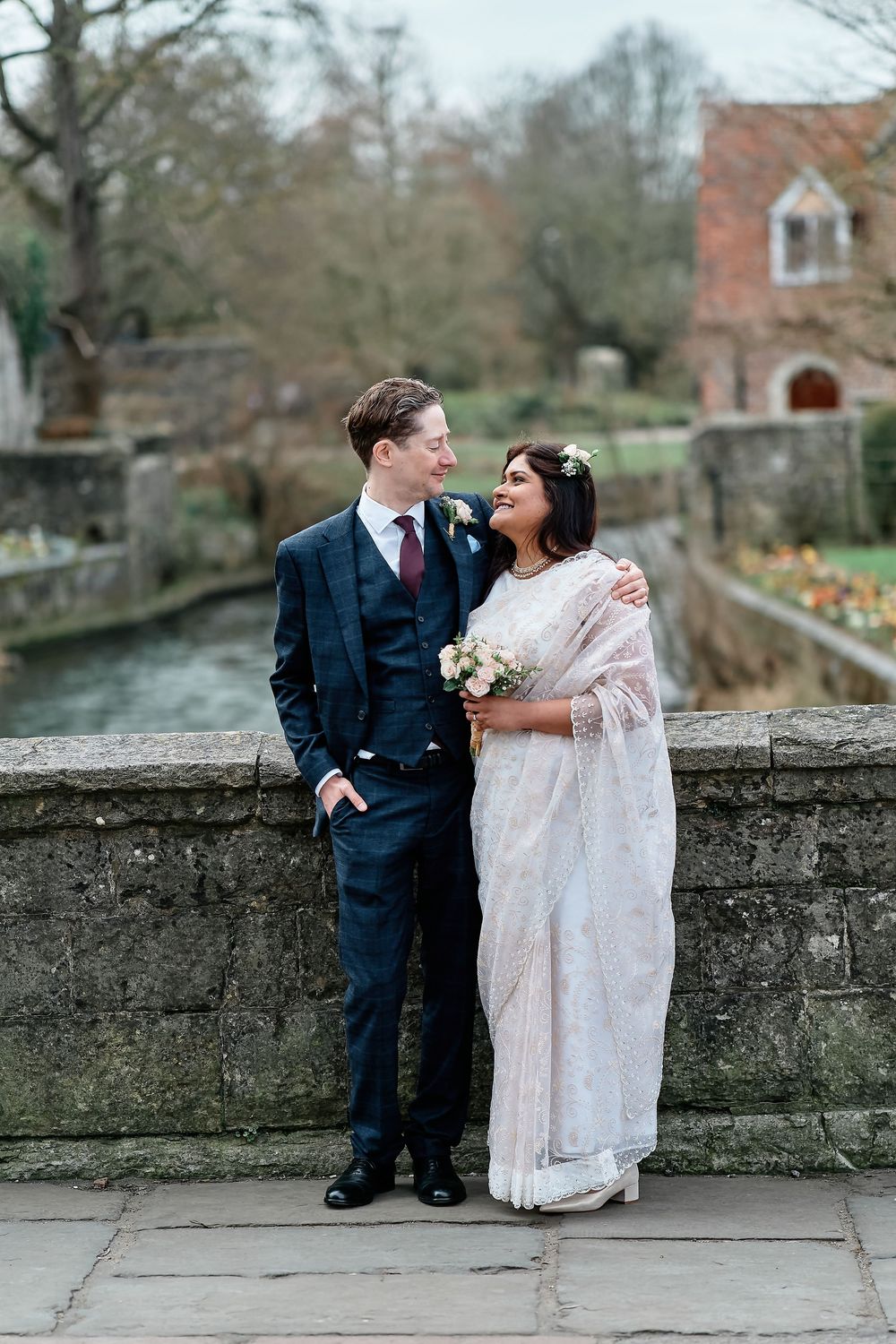 Couple in wedding attire stand together on stone bridge with brick building and garden in background.