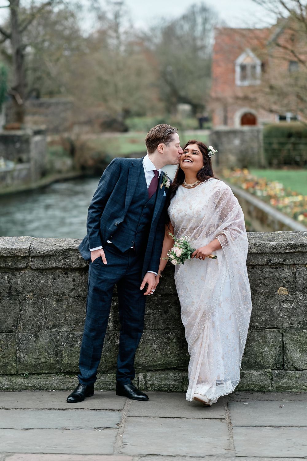 A couple shares a romantic moment on a stone bridge during their wedding day in an English garden setting.