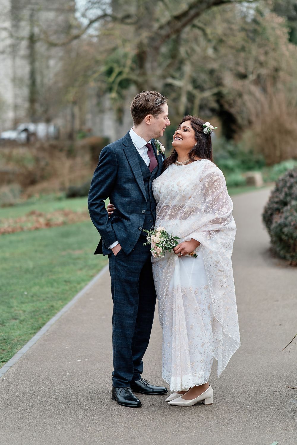 A couple in wedding attire share an intimate moment on a garden path surrounded by trees and greenery.