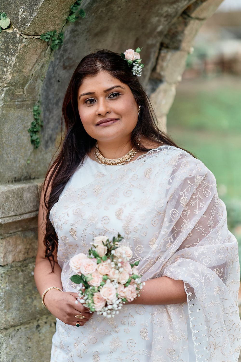 A person wearing a white lace sari holds a small bouquet of pink and white flowers while posing against a stone wall.