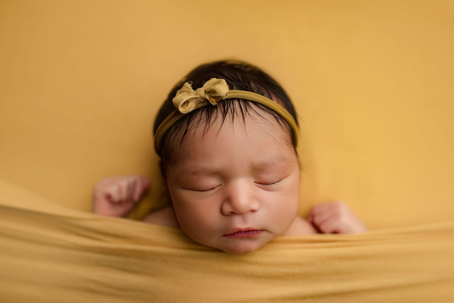 Sleeping newborn wrapped in soft yellow fabric wearing delicate bow headband in peaceful studio portrait.
