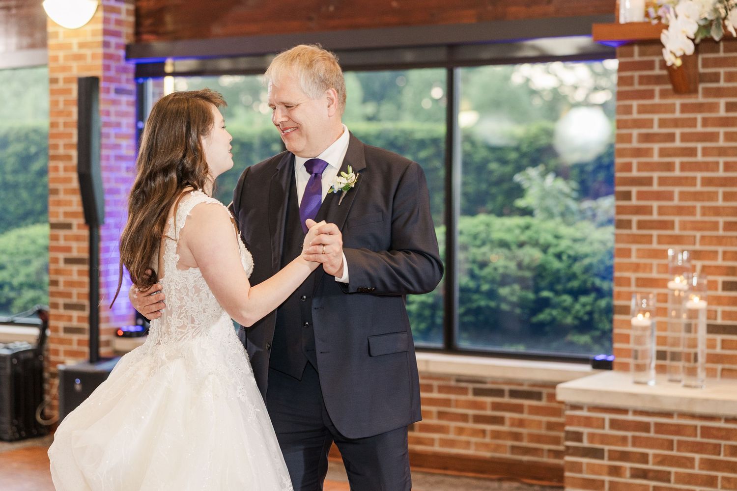 Two people dance together in formal attire at a wedding reception venue with exposed brick walls and large windows.