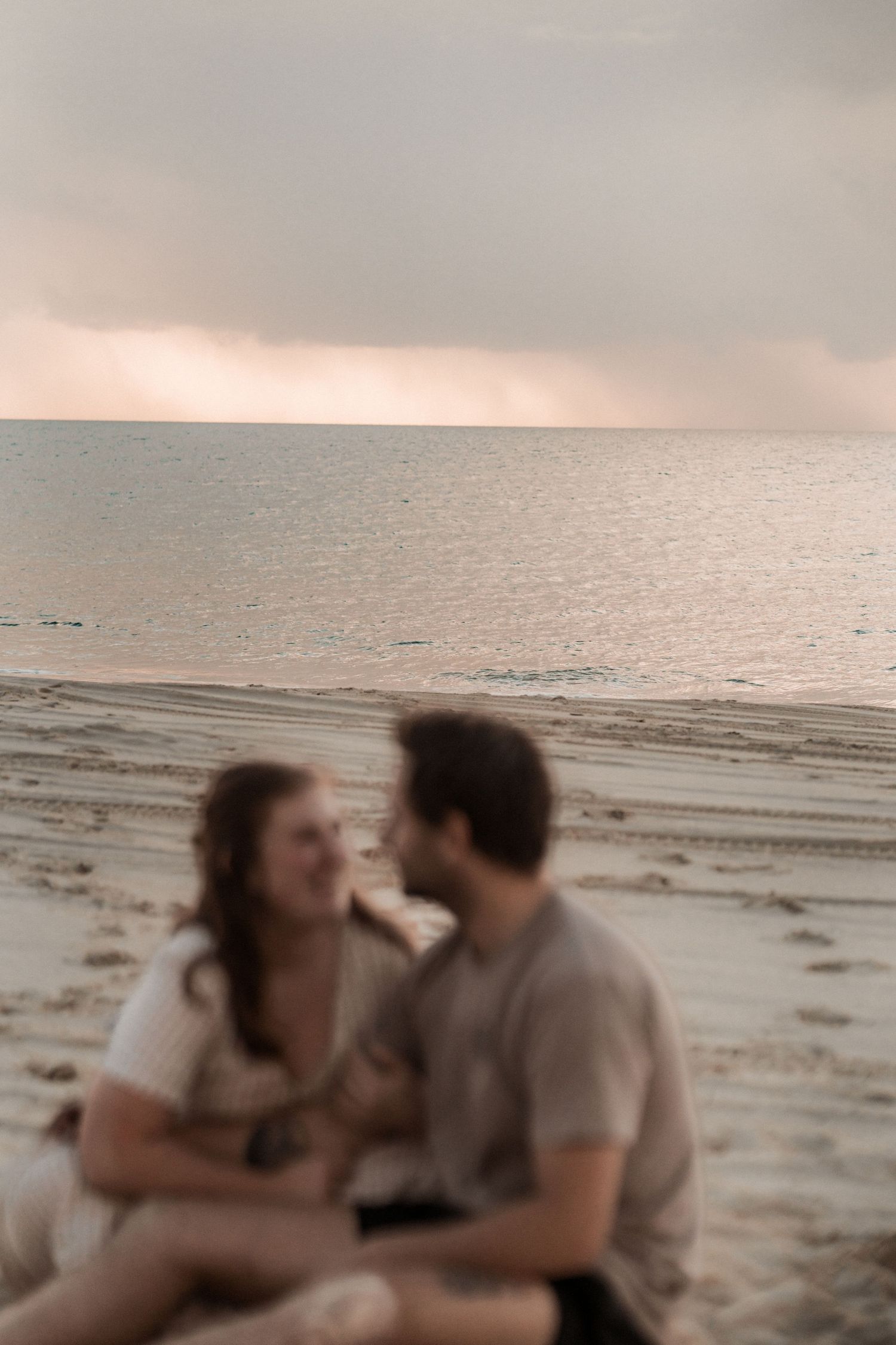 Couple sitting close together on a beach at sunset with soft pink sky over calm ocean waters at Fraser Island, K'gari