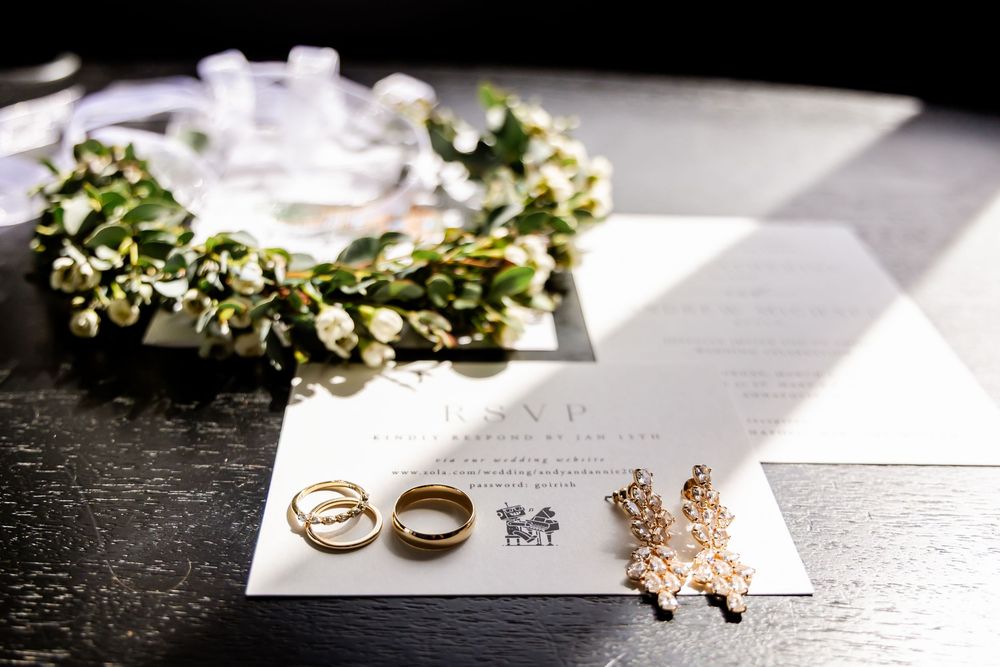 Wedding rings, floral crown, and rose gold jewelry arranged on an invitation card against a dark textured surface.