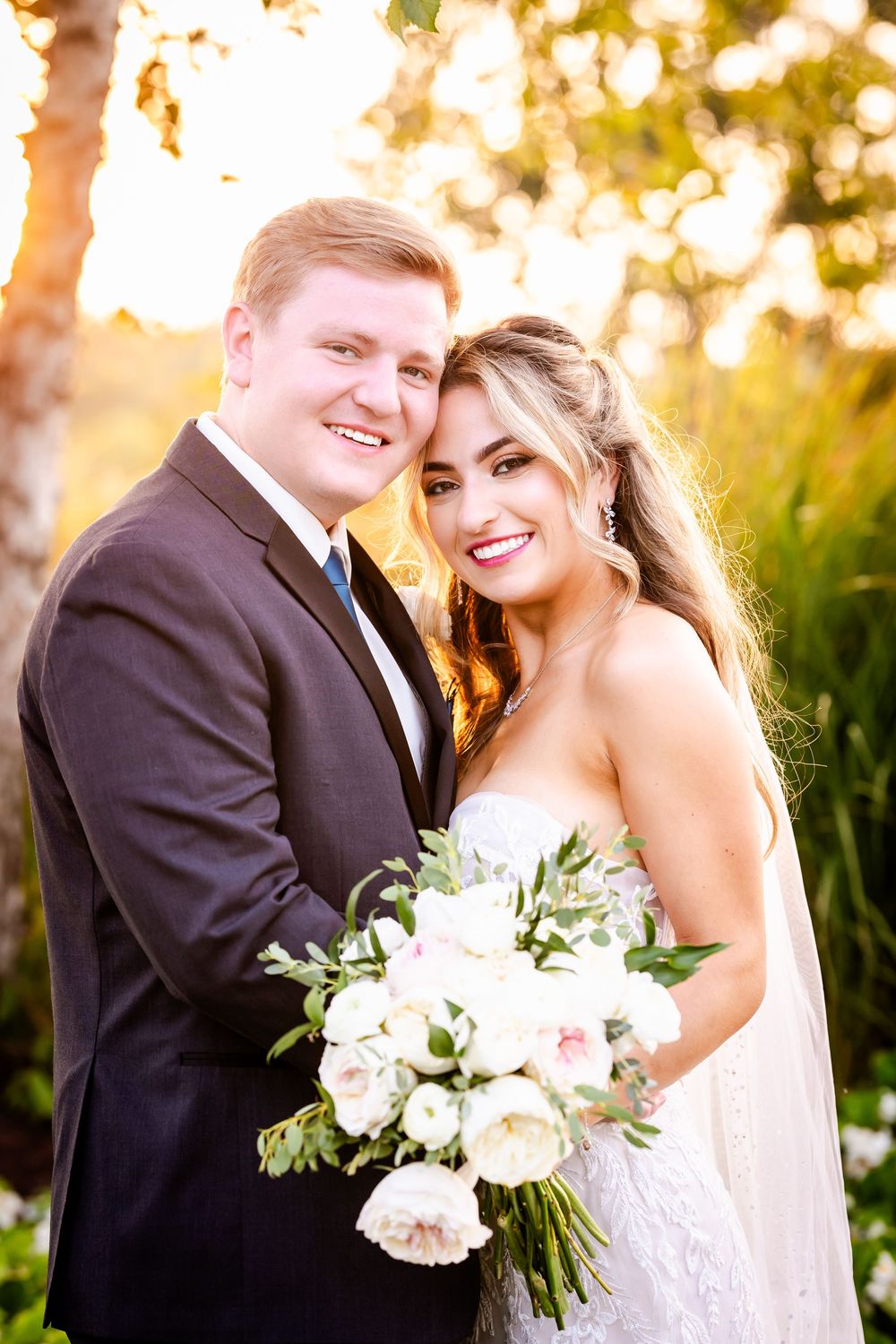 A bride in a white gown holds a blush-colored bouquet while posing with her groom at sunset in a garden setting.