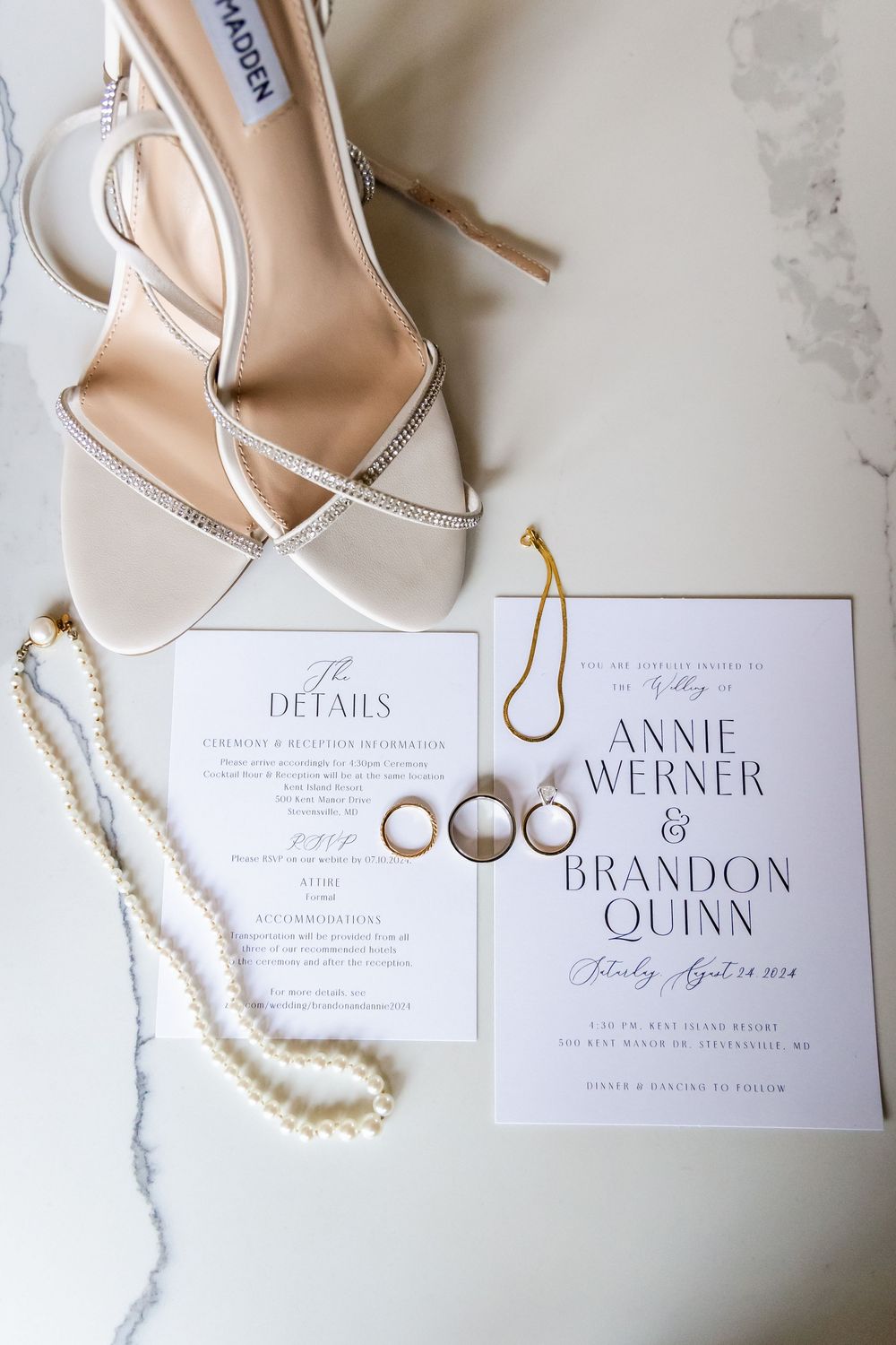 Wedding invitation with elegant beige heels, pearl necklace, and wedding rings on white marble surface.