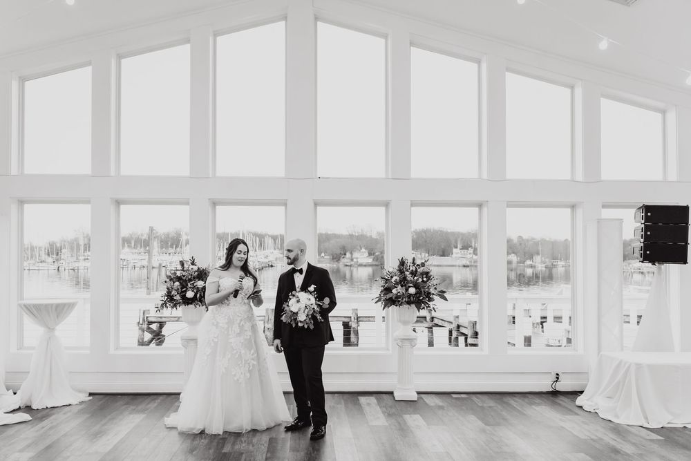 A black and white photograph of a wedding couple standing in an elegant room with large windows.