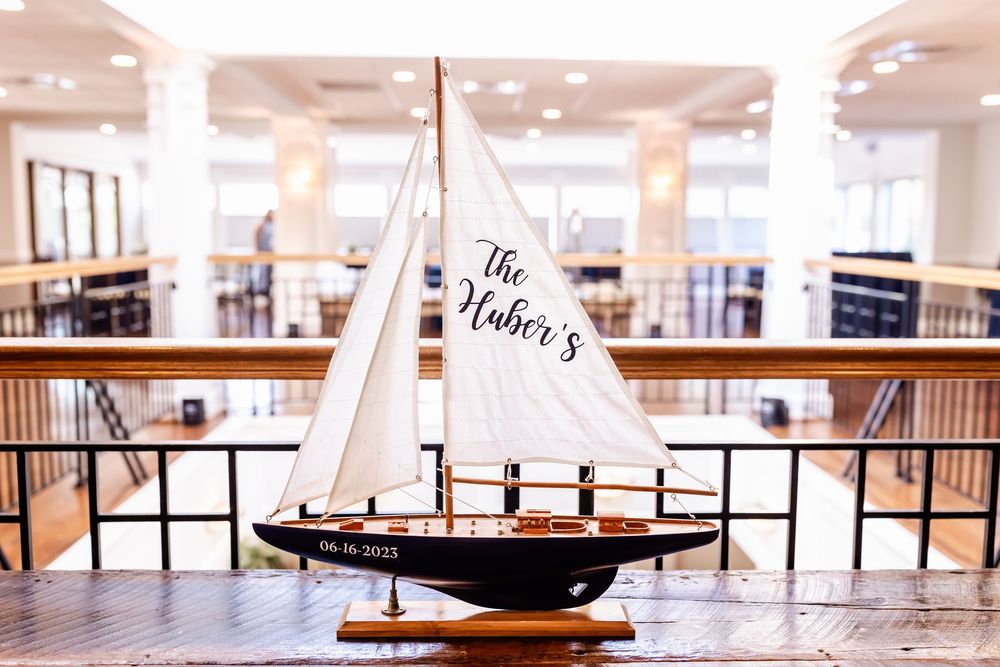 Model sailboat with white sails displayed on wooden table in indoor gallery space with balconies visible in background.